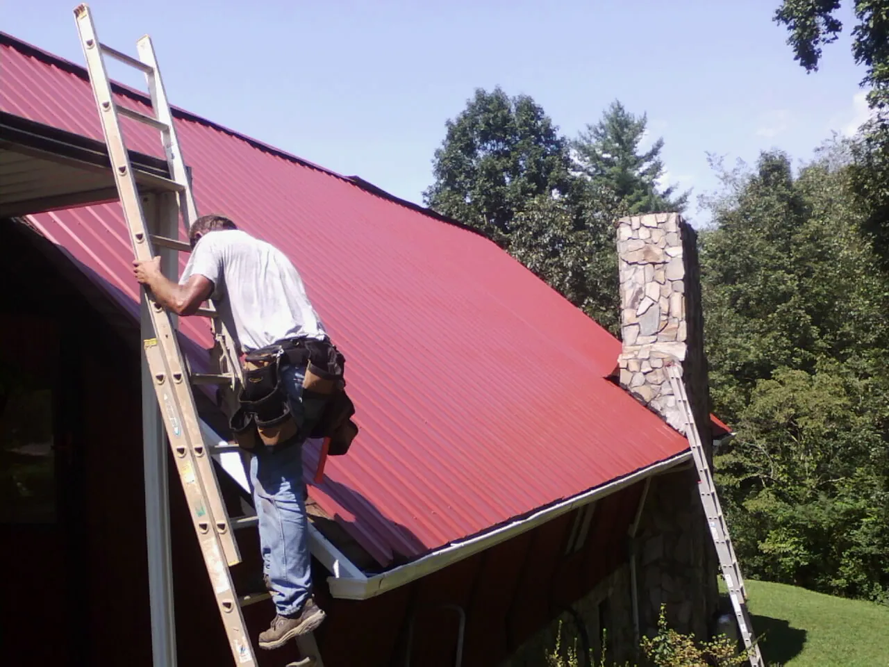 Roofer on a ladder beside a red metal roof and stone chimney — Soffit & Fascia Repair by Roofing Brothers in Nashua